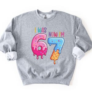 May include: A gray sweatshirt with rolled-up sleeves. The front features the text "I WAS 6, NOW I'M 7" in colorful letters. The numbers 6 and 7 are designed as a pink donut and a blue ice cream cone.