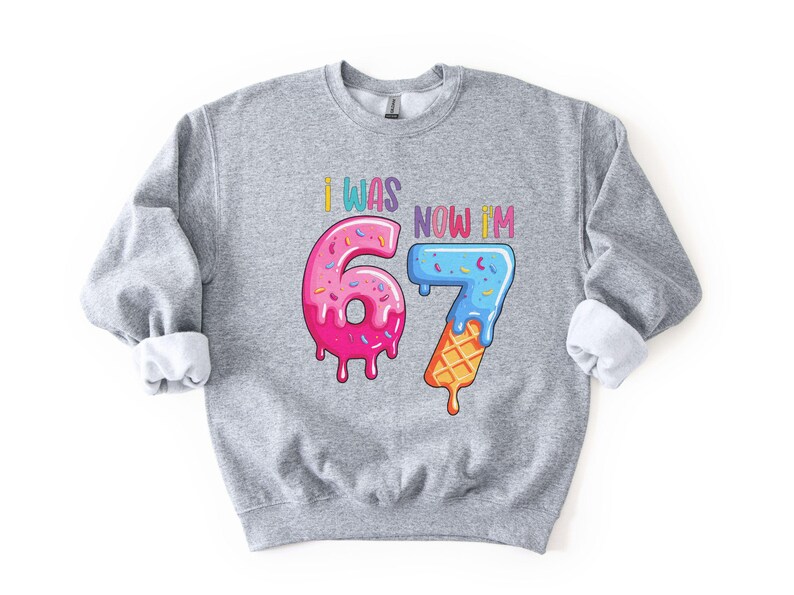 May include: A gray sweatshirt with rolled-up sleeves. The front features the text "I WAS 6, NOW I'M 7" in colorful letters. The numbers 6 and 7 are designed as a pink donut and a blue ice cream cone.