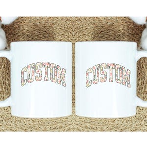 May include: Two white ceramic mugs with a floral pattern spelling out the word "CUSTOM" in a bold, arched font.