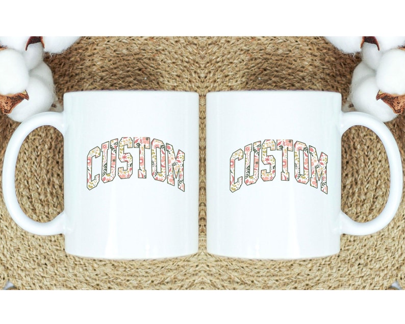 May include: Two white ceramic mugs with a floral pattern spelling out the word "CUSTOM" in a bold, arched font.