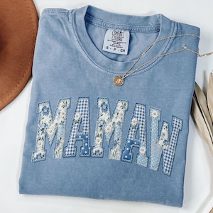 May include: A light blue Comfort Colors t-shirt with the word "MAMAW" in a patchwork design. The letters feature floral, gingham, and polka dot patterns. A gold necklace with a pendant is draped across the shirt.