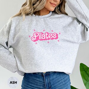 May include: A gray sweatshirt with a pink "Pilates" graphic and stars. The sweatshirt is a relaxed fit and has a crew neck.