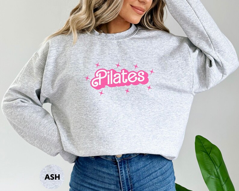May include: A gray sweatshirt with a pink "Pilates" graphic and stars. The sweatshirt is a relaxed fit and has a crew neck.