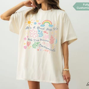 May include: Ivory oversized t-shirt featuring a colourful graphic. The design includes a sun, rainbow, flowers, and the text "It's a Good Day To Help Tiny Humans Communicate". The shirt has a relaxed fit.