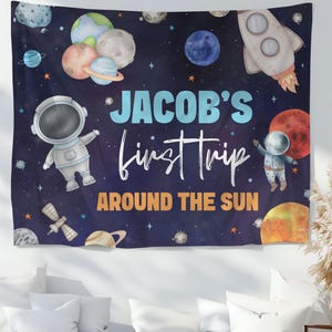 Peut inclure: Une tapisserie bleu marine avec un thème spatial, représentant des planètes, des étoiles et une fusée. Le texte "Jacob's first trip around the sun" est en blanc et orange. Comprend des illustrations d'un astronaute et d'un satellite.