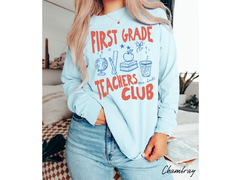 May include: Light blue long-sleeve shirt with the text "FIRST GRADE TEACHERS CLUB" in red. The shirt features school-themed graphics, including a globe, apple, books, and a drink. The name "Mrs. Smith" is also included.