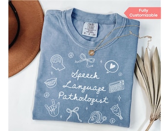 May include: A light blue t-shirt with a white graphic design that says "Speech Language Pathologist". The graphic includes illustrations of a brain, mouth, ear, and hand signing.