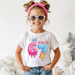 May include: A white t-shirt with the text "I WAS 6, NOW I'M 7" in colorful, playful lettering. The number 6 is designed as a pink donut with sprinkles, and the 7 is an ice cream cone. The girl is wearing sunglasses and a pink headband.