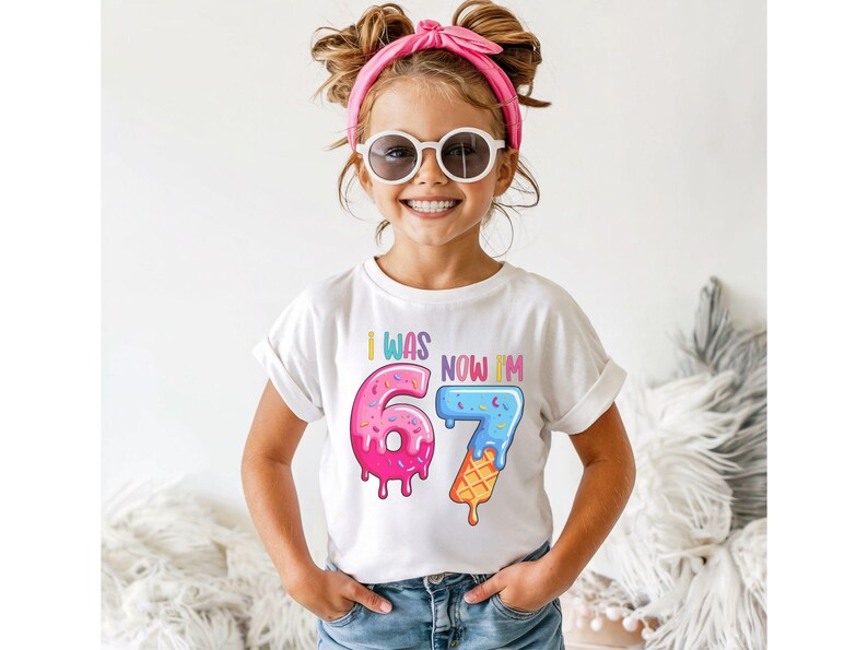 May include: A white t-shirt with the text "I WAS 6, NOW I'M 7" in colorful, playful lettering. The number 6 is designed as a pink donut with sprinkles, and the 7 is an ice cream cone. The girl is wearing sunglasses and a pink headband.