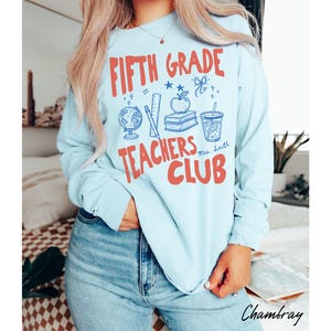 May include: Light blue long-sleeve shirt with the text "FIFTH GRADE TEACHERS CLUB" in red and blue. The design includes a globe, apple, books, and a drink. The shirt is made of a soft material.