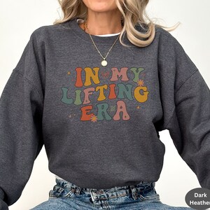 May include: Dark heather gray sweatshirt with the text "IN MY LIFTING ERA" in colorful retro font. The design includes small flower and star accents. A casual, comfortable top for fitness enthusiasts.