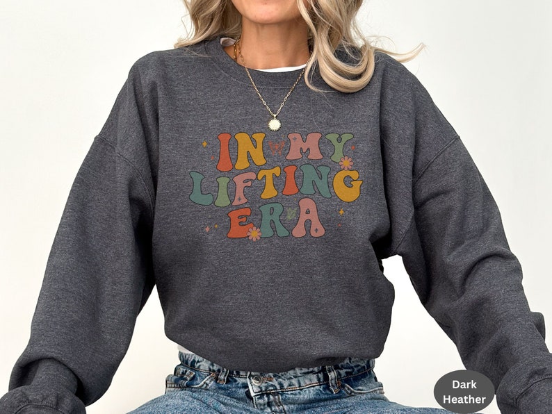 May include: Dark heather gray sweatshirt with the text "IN MY LIFTING ERA" in colorful retro font. The design includes small flower and star accents. A casual, comfortable top for fitness enthusiasts.
