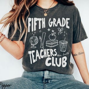 May include: Dark gray t-shirt with the words "FIFTH GRADE TEACHERS CLUB" in white, along with illustrations of a globe, apple, books, and a drink. The shirt has a vintage, worn look.