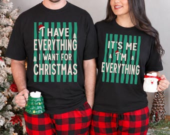 May include: Two black t-shirts featuring green vertical stripes and white text. The left shirt reads I HAVE EVERYTHING I WANT FOR CHRISTMAS with a small Santa hat and tree. The right shirt reads IT'S ME I'M EVERYTHING with a Santa hat and tree.
