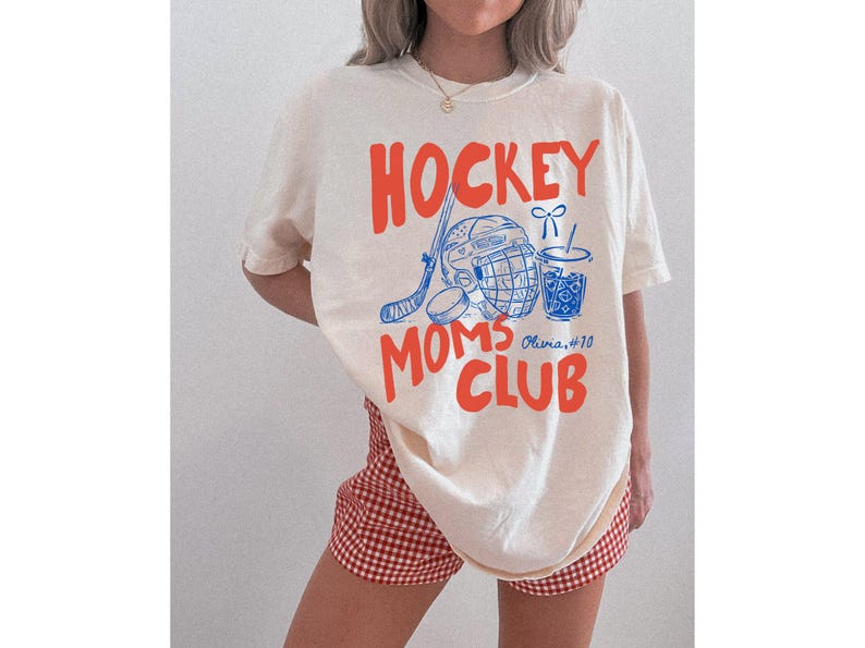 May include: Cream-colored t-shirt with the words "HOCKEY MOMS CLUB" in orange, along with blue hockey-themed graphics. The shirt also features the name "Olivia #10". The person is wearing red and white checkered shorts.