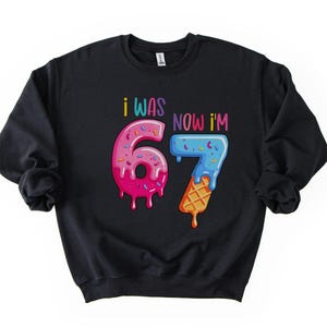 May include: Black sweatshirt with the text "I WAS 67 NOW I'M 67" in colorful, playful lettering. The numbers are designed to look like a donut and an ice cream cone, with dripping frosting and sprinkles.