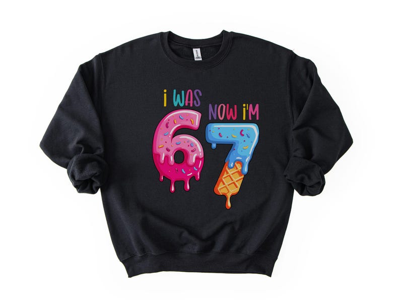 May include: Black sweatshirt with the text "I WAS 67 NOW I'M 67" in colorful, playful lettering. The numbers are designed to look like a donut and an ice cream cone, with dripping frosting and sprinkles.