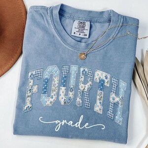 May include: A light blue Comfort Colors t-shirt with the words "FOURTH grade" in a patchwork design. The letters are in various blue floral and gingham patterns. A gold necklace with a pendant rests on the shirt.