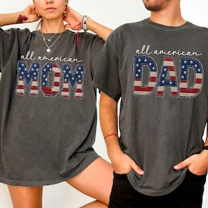 May include: Two charcoal gray t-shirts. One says "all american MOM" and the other says "all american DAD". The text is in white script, with the letters MOM and DAD filled with a red, white, and blue American flag design.