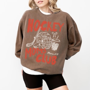 May include: Brown sweatshirt with the text "HOCKEY MOMS CLUB" in red. The design includes a hockey stick, helmet, puck, and a drink with a straw. The sweatshirt has a vintage look.