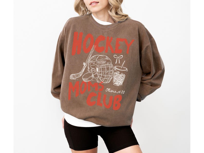 May include: Brown sweatshirt with the text "HOCKEY MOMS CLUB" in red. The design includes a hockey stick, helmet, puck, and a drink with a straw. The sweatshirt has a vintage look.