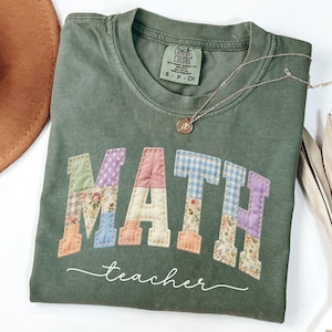 May include: A moss green t-shirt with the word "MATH" in patchwork letters, and the word "teacher" in a cursive font. A gold necklace with a pendant is on the shirt. A brown hat and dried plants are in the background.