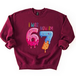 May include: Burgundy sweatshirt with the text "I WAS 6 NOW I'M 7". The numbers are designed as donuts and ice cream cones with colorful sprinkles and dripping icing. A fun birthday-themed graphic.