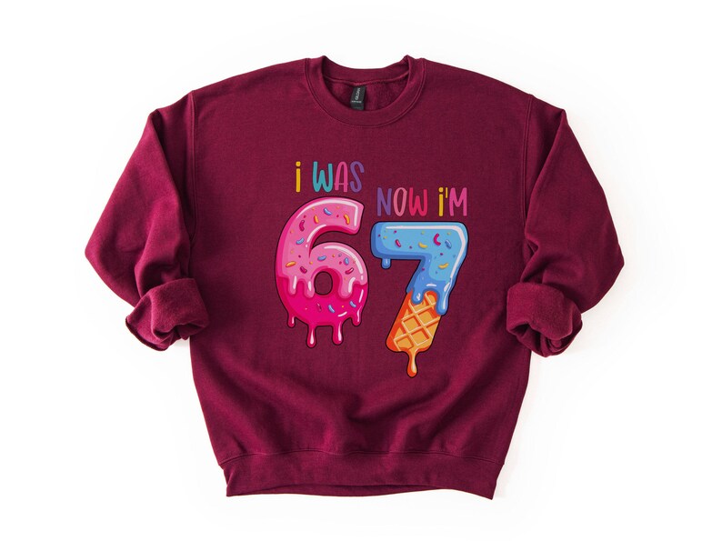 May include: Burgundy sweatshirt with the text "I WAS 6 NOW I'M 7". The numbers are designed as donuts and ice cream cones with colorful sprinkles and dripping icing. A fun birthday-themed graphic.