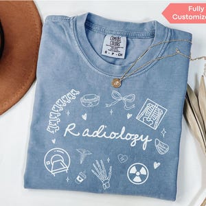 Op de afbeelding: Een lichtblauw T-shirt met een wit grafisch ontwerp met het woord "Radiologie" en verschillende medische symbolen, waaronder een röntgenfoto, een stethoscoop en een bot. Het shirt is gemaakt van een zacht, comfortabel materiaal en is volledig aanpasbaar.