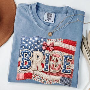 May include: A light blue Comfort Colors t-shirt featuring a patchwork American flag design with the word "BRIDE" in a decorative font. A gold necklace and a brown hat are also visible.