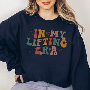 May include: Navy blue sweatshirt with the text "IN MY LIFTING ERA" in retro-style lettering. The design includes butterflies and flowers in shades of orange, yellow, green, and pink. A casual, comfortable top for fitness enthusiasts.