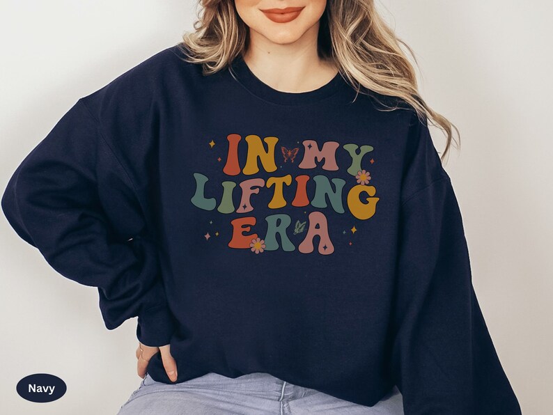 May include: Navy blue sweatshirt with the text "IN MY LIFTING ERA" in retro-style lettering. The design includes butterflies and flowers in shades of orange, yellow, green, and pink. A casual, comfortable top for fitness enthusiasts.