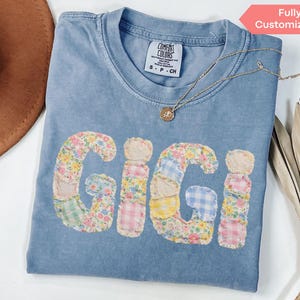 May include: A light blue Comfort Colors t-shirt with the name "GIGI" appliqued in a patchwork floral design. A gold necklace with a pendant rests on the shirt. The words "Fully Customizable" are in a pink banner.