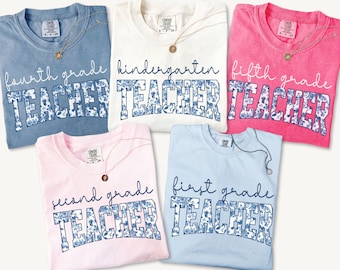 May include: Five t-shirts in various colors: light blue, white, pink, light pink, and hot pink. Each shirt has a grade level and the word "TEACHER" in a floral design. A gold necklace with a pendant is on each shirt.