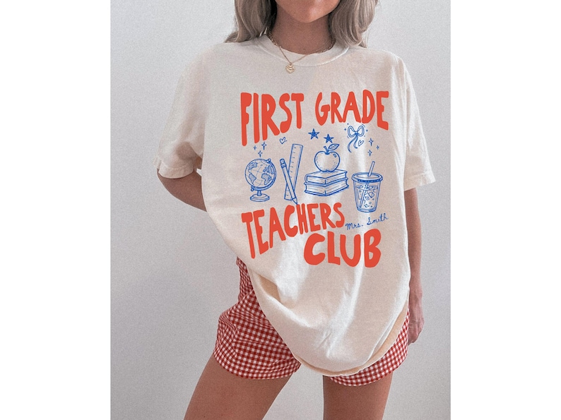 Woman wearing an oversized ivory First Grade Teachers Club shirt with school themed graphics including books, apple, globe, ruler, and drink. This personalized teacher tee is a cute choice for back to school, classroom wear, and teacher team outfits.