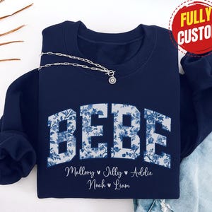 May include: Navy blue sweatshirt with the word "BEBE" in a floral design. Below the word are the names Mallory, Jilly, Addie, Noah, and Liam. A silver necklace with a pendant is draped across the sweatshirt. A red circle with the words "FULLY CUSTOM" is in the upper right corner.