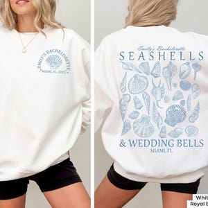 May include: White sweatshirt with a blue seashell design on the back and the text "Emily's Bachelorette Seashells & Wedding Bells Miami, FL" printed on the back. The front of the sweatshirt has the text "Emily's Bachelorette Miami, FL 2025" printed in blue.