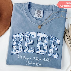 May include: A blue jean Comfort Colors t-shirt with the word "BEBE" in large, floral-patterned letters. Below the word are names: Mallory, Jilly, Addie, Noah, and Liam. A gold necklace and a brown hat are also visible.