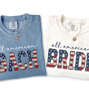 May include: Two t-shirts, one light blue and one white, with the words "all american BACH" and "all american BRIDE" in a patriotic font. Each shirt has a gold necklace.
