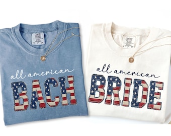 Könnte beinhalten: Zwei T-Shirts, eines hellblau und eines weiß, mit den Worten "all american BACH" und "all american BRIDE" in einer patriotischen Schriftart. Jedes Shirt hat eine goldene Halskette.