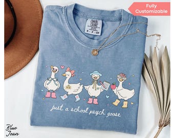 May include: A blue t-shirt featuring a cartoon goose design with the text "just a school psych goose". The geese are wearing accessories and boots. A gold necklace is draped on the shirt. The t-shirt is a blue jean color.