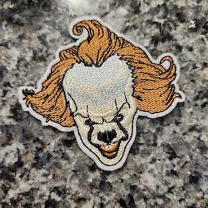 May include: Embroidered patch of a clown's face with a menacing grin. The clown has orange hair, white skin, and red and black makeup around the eyes and mouth.