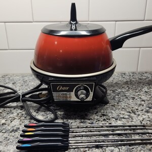 May include: An electric Oster fondue pot with a red bowl, black lid, and black handle. The base features a temperature control dial with the text Oster Electric Fondue. Six metal fondue forks with colored ends are arranged in front of the unit.