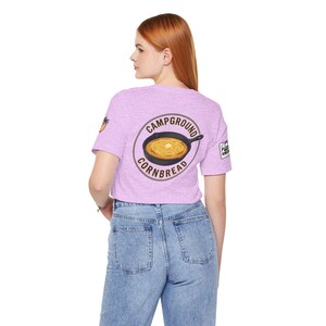 May include: A light purple crop top with a graphic of cornbread in a skillet, encircled by the words "Campground Cornbread." The shirt also features a small palm tree graphic and a white square logo.