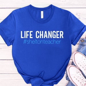 May include: A royal blue t-shirt with the text "LIFE CHANGER #sheltonteacher" in white and light blue.