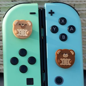 May include: A pair of Nintendo Switch Joy-Con controllers with custom bear-shaped button caps. The left Joy-Con is mint green and the right Joy-Con is light blue.