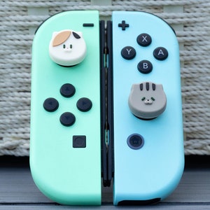 May include: A turquoise and light blue Nintendo Switch controller with cat-shaped joy-con caps. The left joy-con has a white cat face with brown ears and the right joy-con has a grey cat face with black ears.