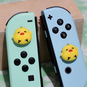 May include: A pair of Nintendo Switch Joy-Cons in pastel shades. One is mint green, the other is light blue. Each has a yellow chick-shaped charm. The mint green Joy-Con has black buttons. The light blue Joy-Con has black buttons and a black joystick.