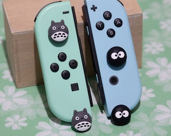 May include: Two Nintendo Switch Joy-Con controllers with custom face buttons. The left controller is light green with a black Totoro character face button. The right controller is light blue with a black No Face character face button.