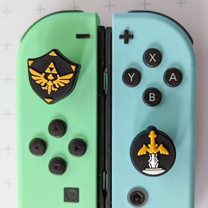 May include: A pair of Nintendo Switch Joy-Con controllers in mint green and light blue. Each controller has black buttons and a decorative emblem with a gold and black design. The controllers are designed with a Legend of Zelda theme.